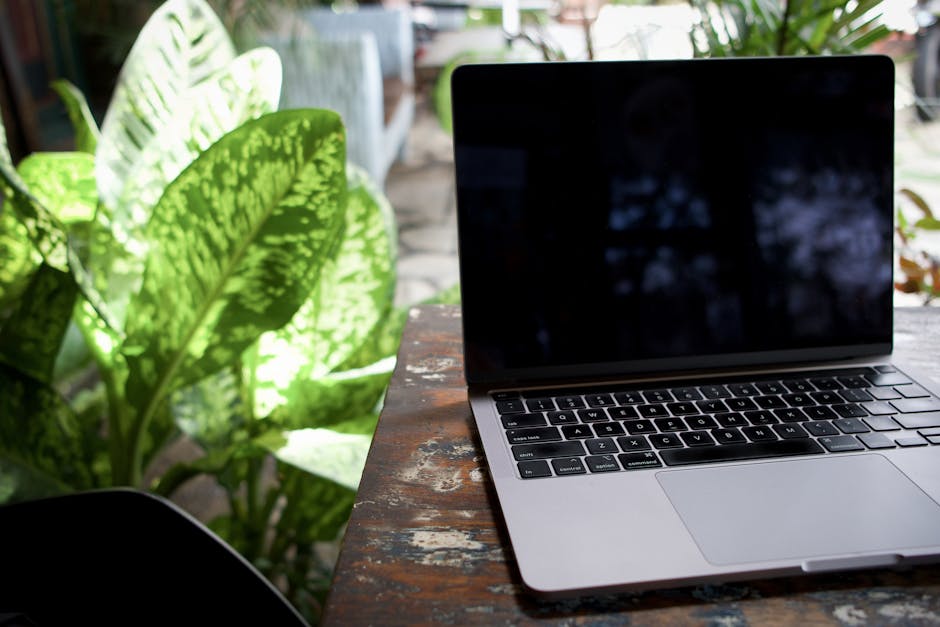 A laptop sits on a rustic table surrounded by lush green plants in an outdoor setting.