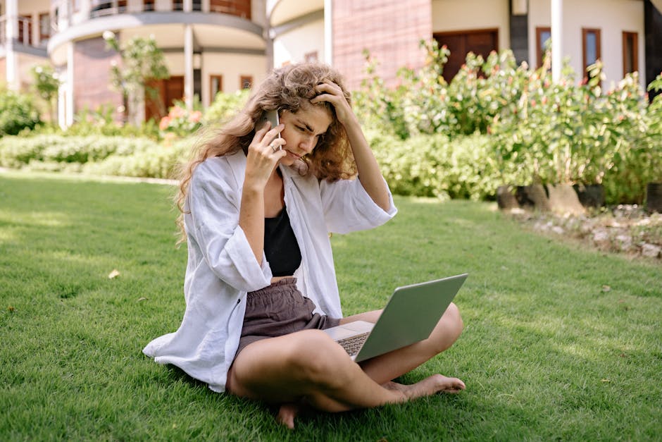 A woman sits on a lawn, using a laptop and phone, working remotely outdoors in a tropical setting.